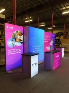 Modified VK-2993 Backlit Modular Exhibit with Tension Fabric Graphics, Monitor Mount, (2) MOD-1701 Backlit Counters with Storage and (1) MOD-1700 Backlit Reception Counter with Wireless Charging Pads